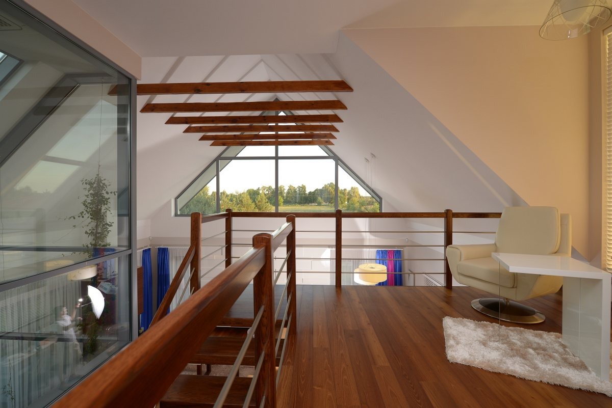 Jasne poddasze z drewnianą balustradą i widokiem na zieleń przez okno. Kremowy fotel i biały stolik na drewnianej podłodze. Elementy dekoracyjne i lampa wisząca.