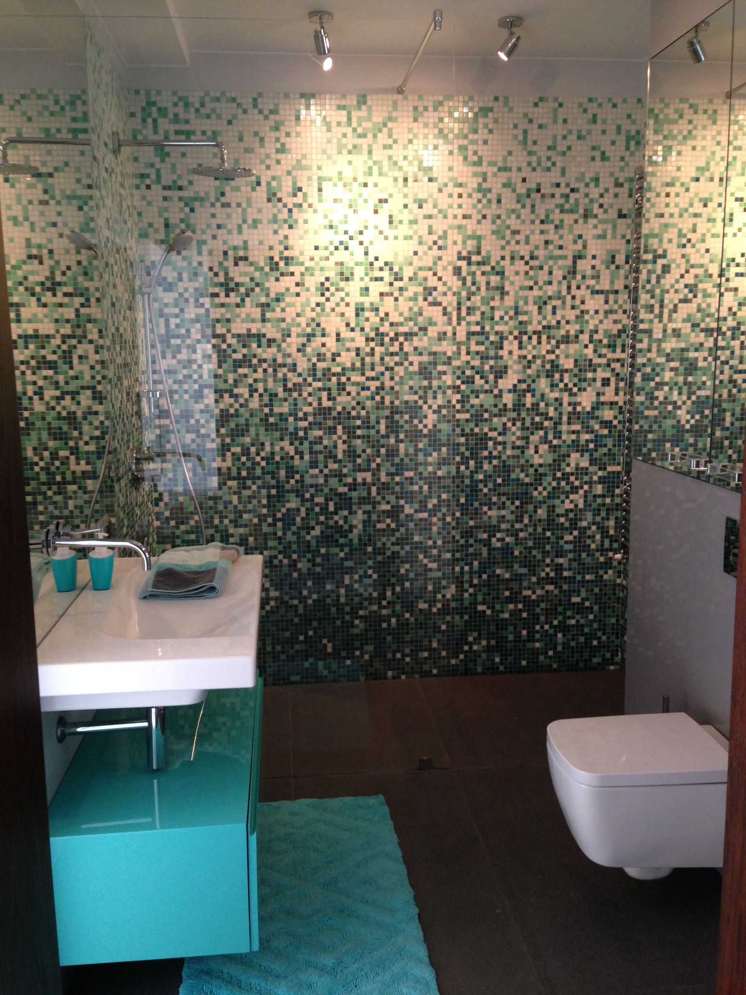 Nowoczesna łazienka z mozaiką w odcieniach zieleni i turkusu. Umywalka z szafką w kolorze turkusowym, biała toaleta podwieszana, brązowe płytki podłogowe.