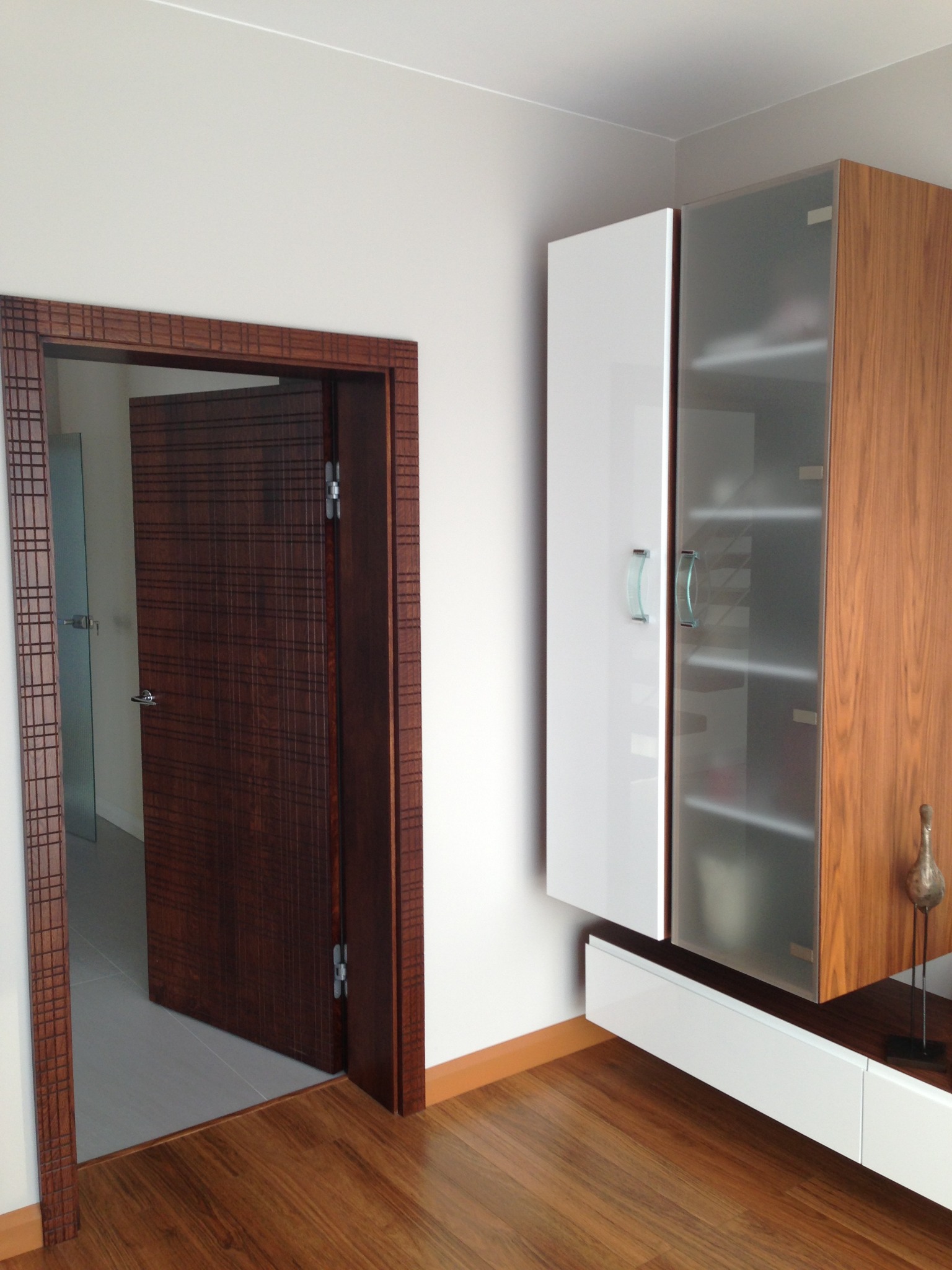 Nowoczesne wnętrze: drewniana podłoga, szafka wisząca z białymi frontami i szklanymi drzwiczkami oraz fornirowane drzwi z ozdobną ościeżnicą.