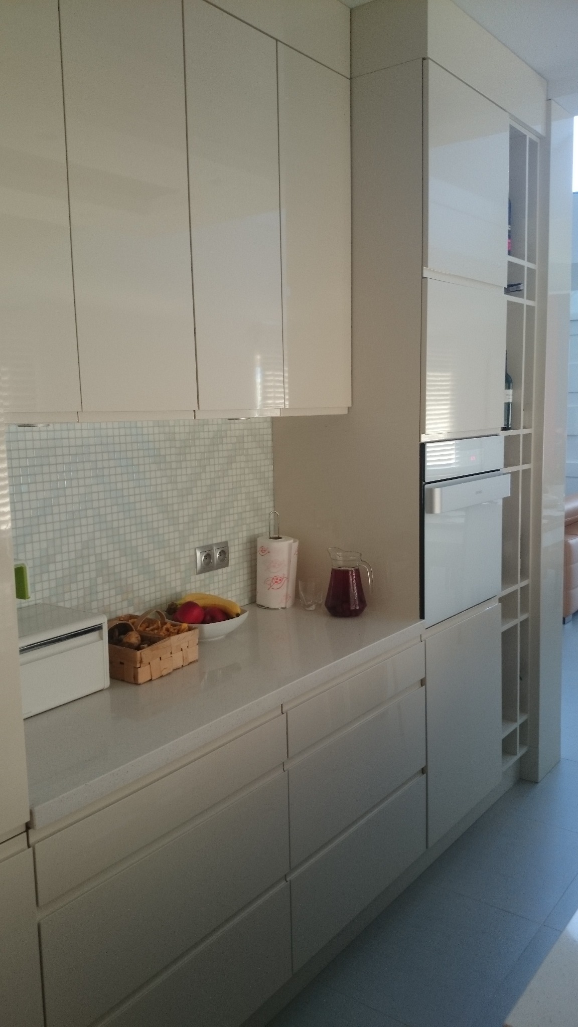 Biała, lśniąca kuchnia z mozaiką nad blatem, owocami i dzbankiem z napojem. Nowoczesny design, jasne oświetlenie, minimalistyczny styl, widoczne gniazdka elektryczne.