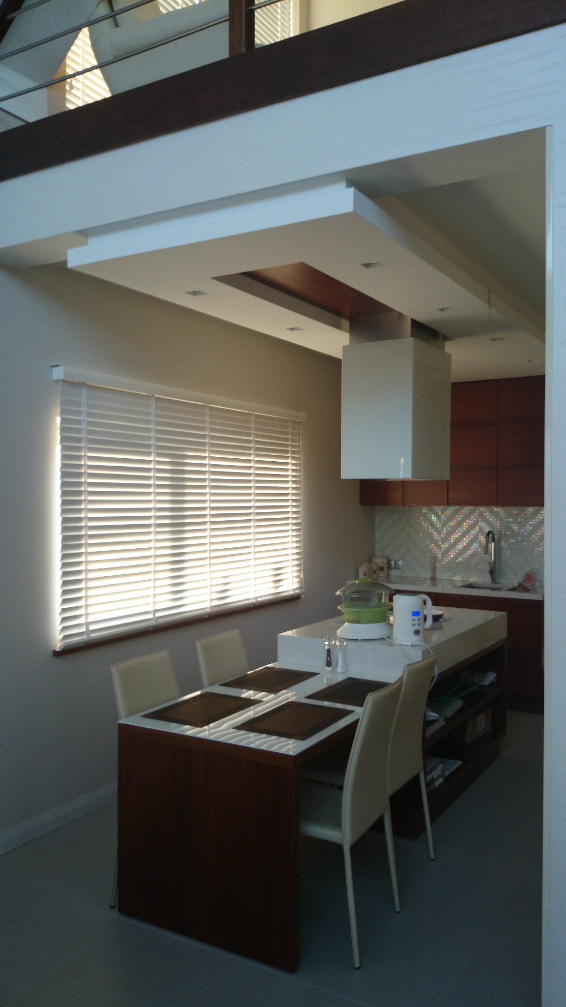 Nowoczesna kuchnia z wyspą i stołem jadalnym zintegrowanym z wyspą, jasne blaty, drewniane akcenty, rolety w oknie, minimalistyczny okap.