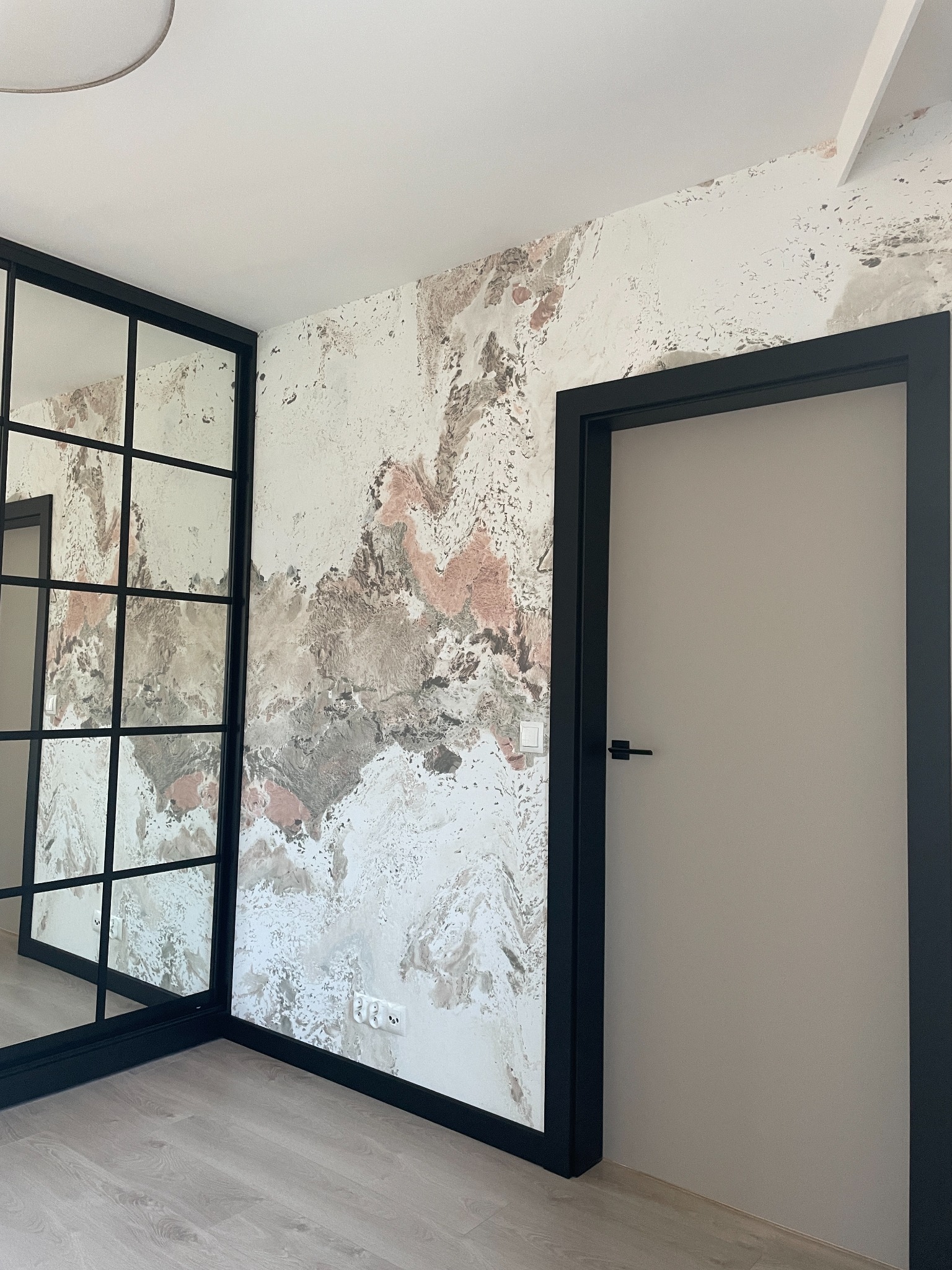 Pokój z dekoracyjną ścianą w odcieniach bieli, szarości i różu, czarną ramą lustra i drzwiami z czarną ościeżnicą. Nowoczesny design.