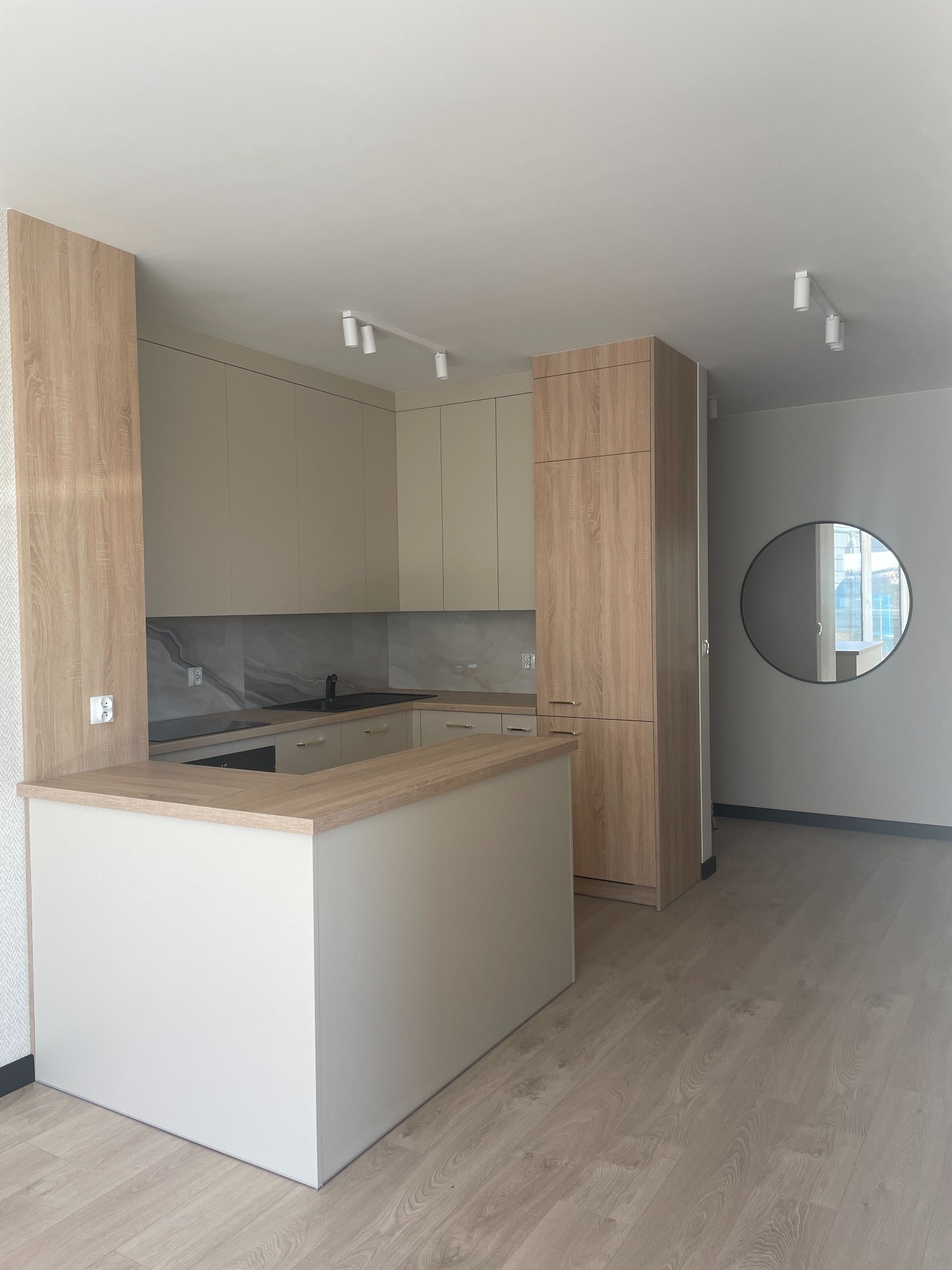 Jasna kuchnia z wyspą, drewniane fronty szafek i okrągłe lustro na ścianie. Minimalistyczne oświetlenie punktowe. Nowoczesny design.