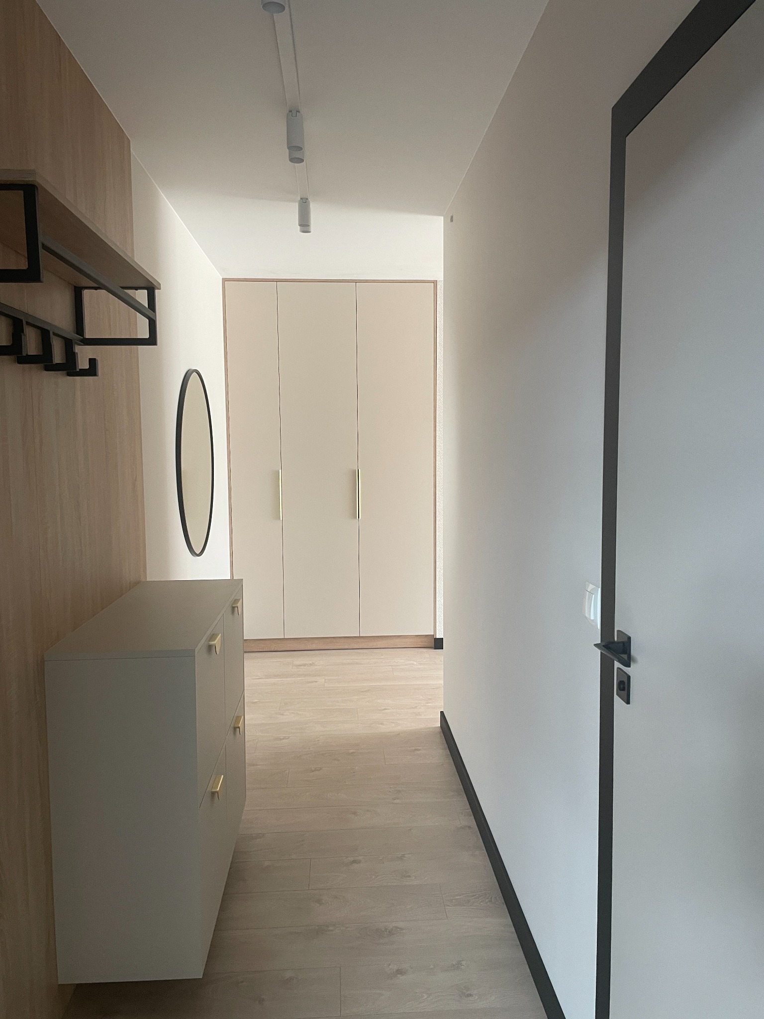 Nowoczesny przedpokój w jasnych barwach: szafa w zabudowie, komoda z złotymi uchwytami, okrągłe lustro i drewnopodobna okładzina ścienna. Minimalistyczny design.
