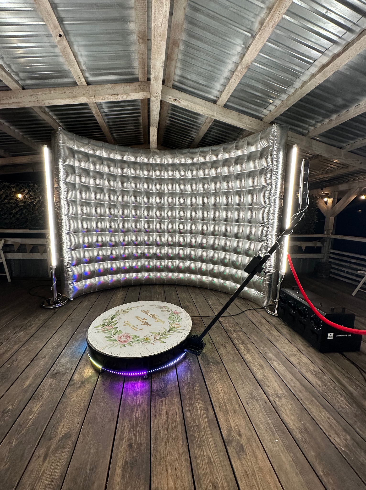Srebrna, dmuchana fotobudka z oświetleniem LED i obrotową platformą na drewnianej podłodze pod zadaszeniem, gotowa do użycia na imprezie.