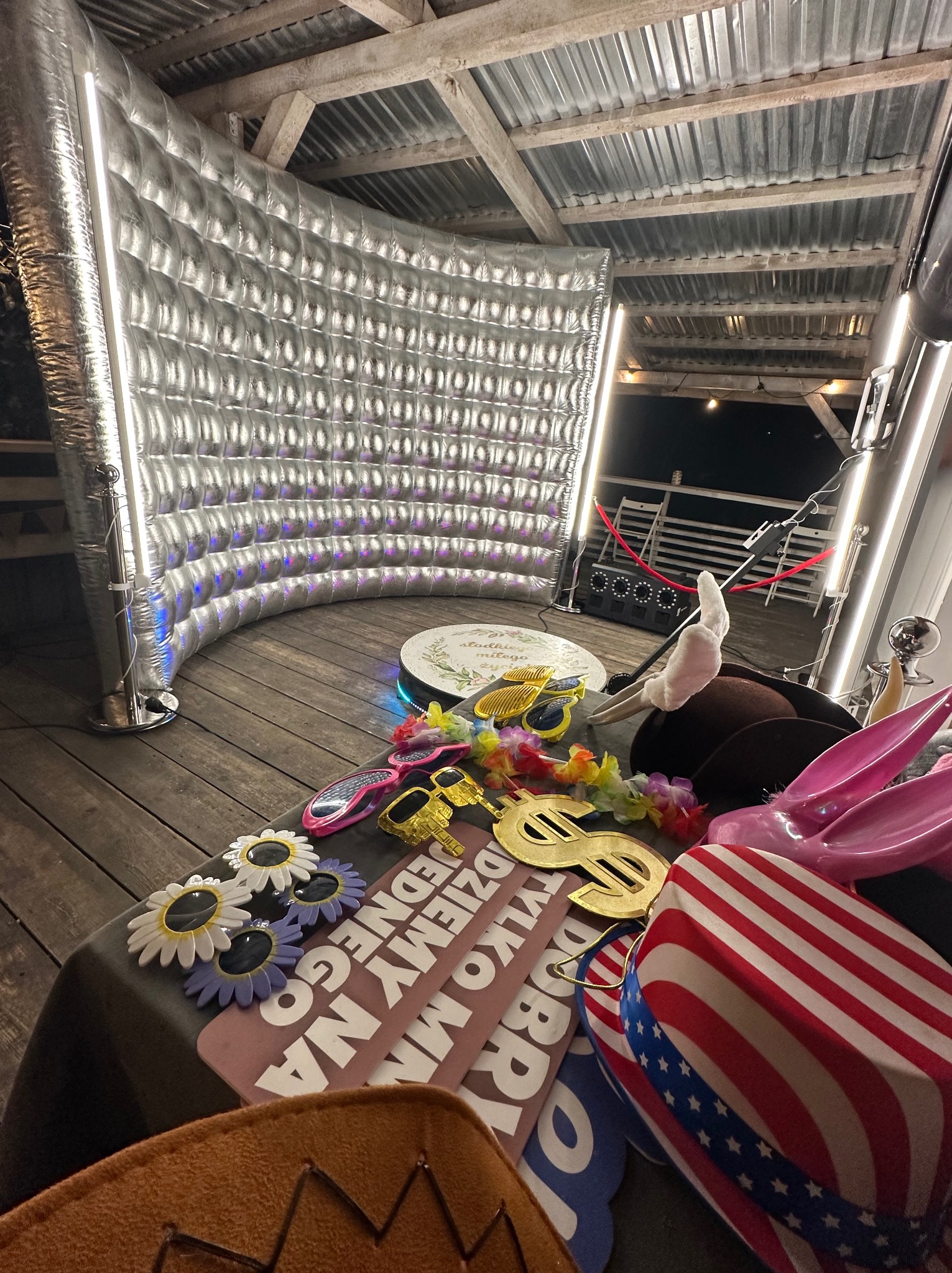 Srebrna fotobudka z rekwizytami: okulary, kapelusze, zabawne napisy. Scena gotowa do sesji zdjęciowej na imprezie plenerowej z drewnianą podłogą i zadaszeniem.