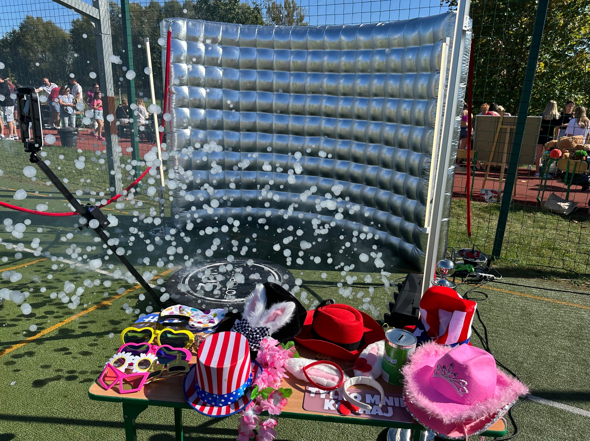 Inscenizacja imprezowa: srebrna ścianka, rekwizyty (kapelusze, okulary) na stole, wytwornica piany w akcji, zielona trawa i ludzie w tle. Zabawa na świeżym powietrzu.