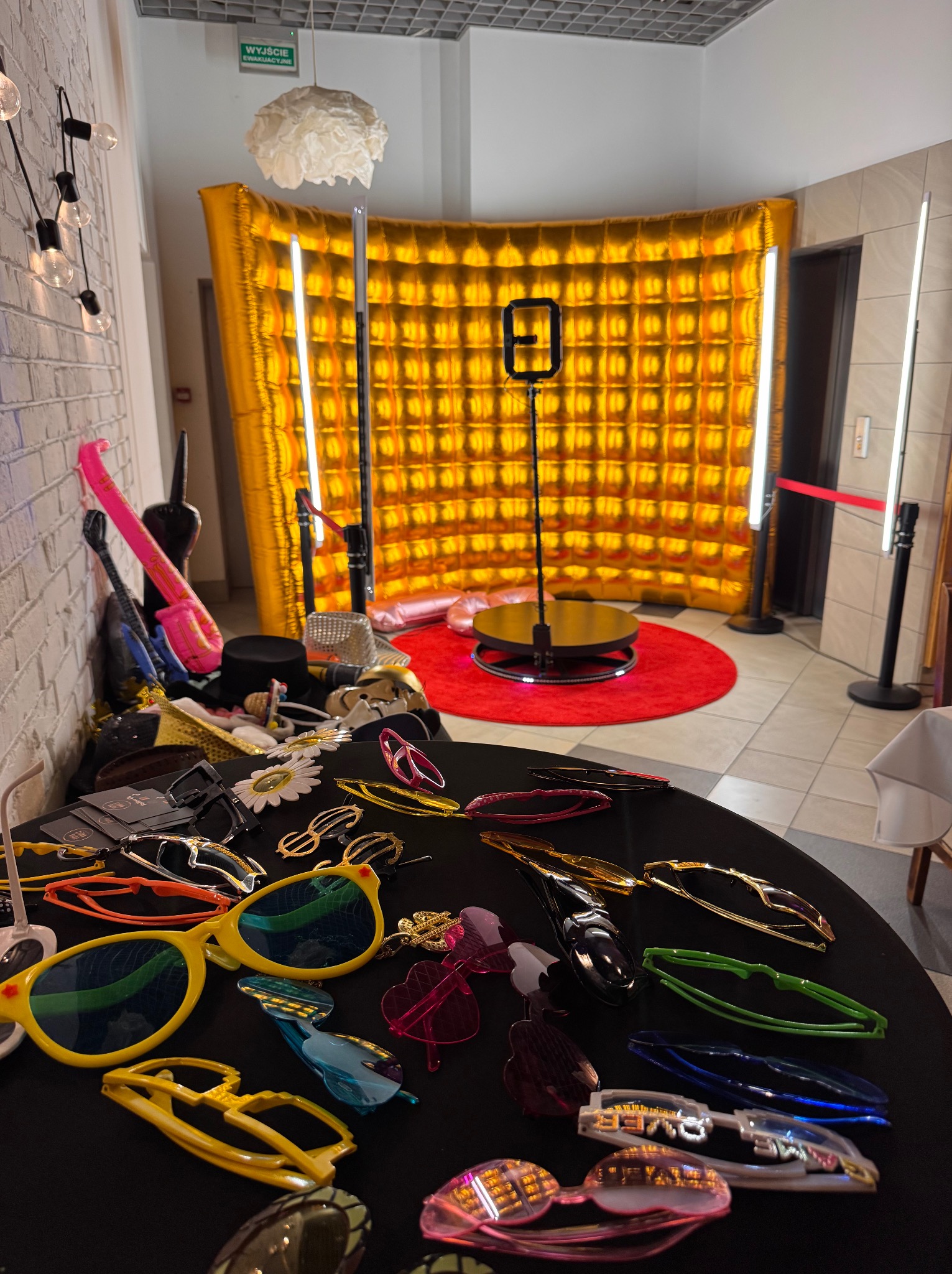 Zestaw rekwizytów (okulary, kapelusze) na stole przed złotą, dmuchaną ścianką fotobudki z podświetleniem LED i czerwonym dywanem. W tle winda.