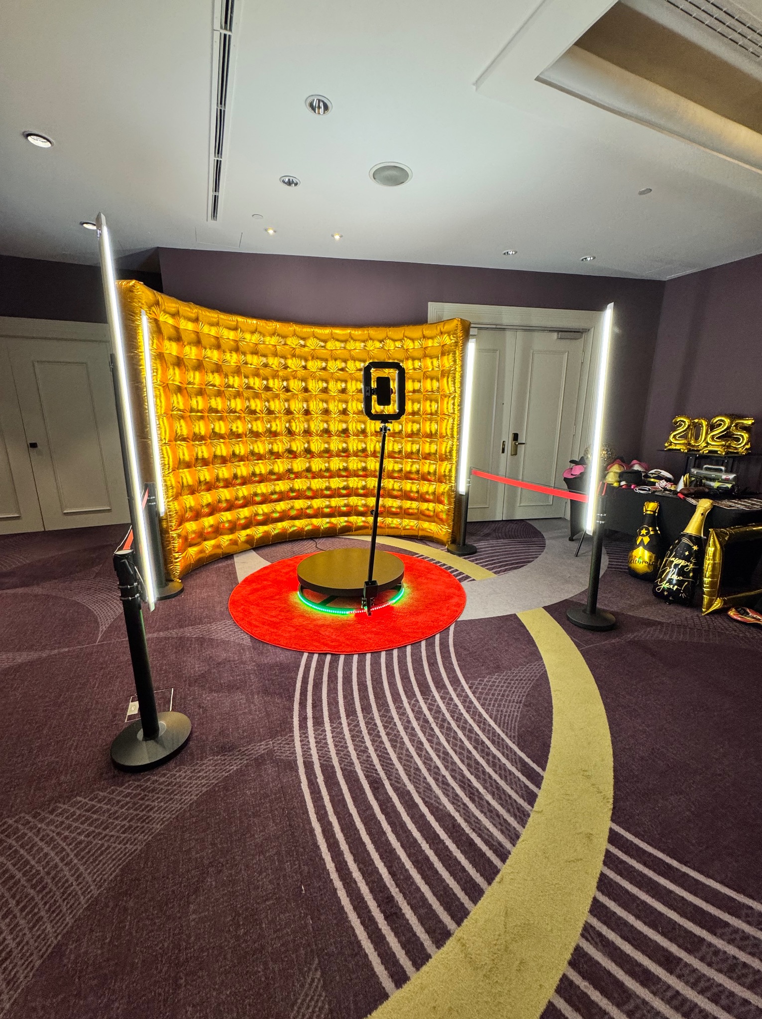 Złota ścianka dmuchana, czerwony dywan i obrotowa platforma z oświetleniem LED tworzą nowoczesną fotobudkę na tle wzorzystej wykładziny i dekoracji '2025'.