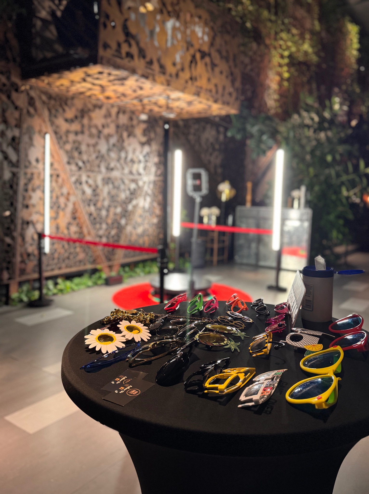 Dekoracyjna fotobudka z rekwizytami: okulary, kwiaty na stole z czarnym obrusem. W tle rustykalna ścianka i czerwony dywan. Oświetlenie ledowe.