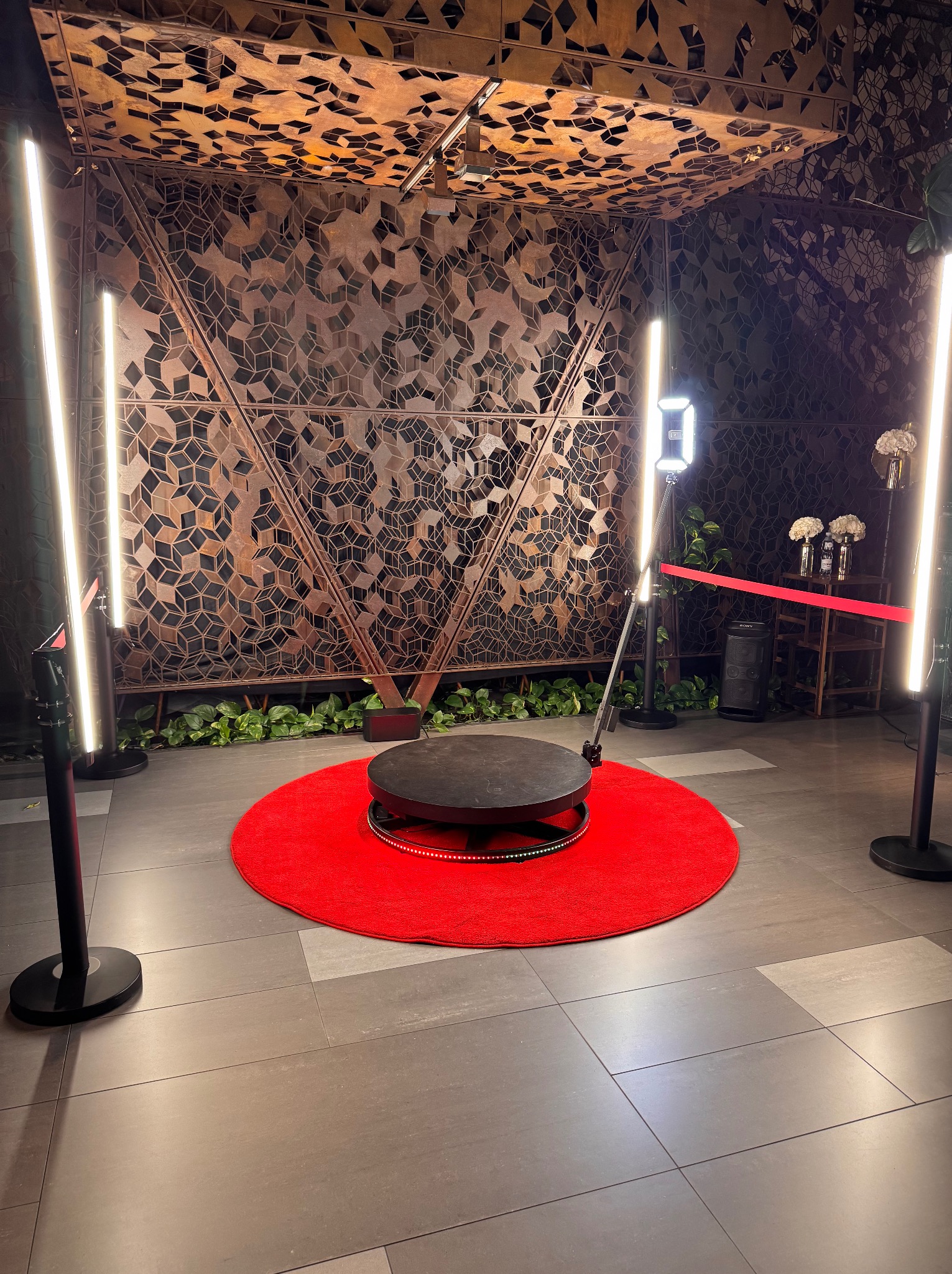 Rotacyjna platforma 360° na czerwonym dywanie, otoczona lampami LED, na tle dekoracyjnej ściany z geometrycznym wzorem, tworząca scenę do nagrywania filmów.