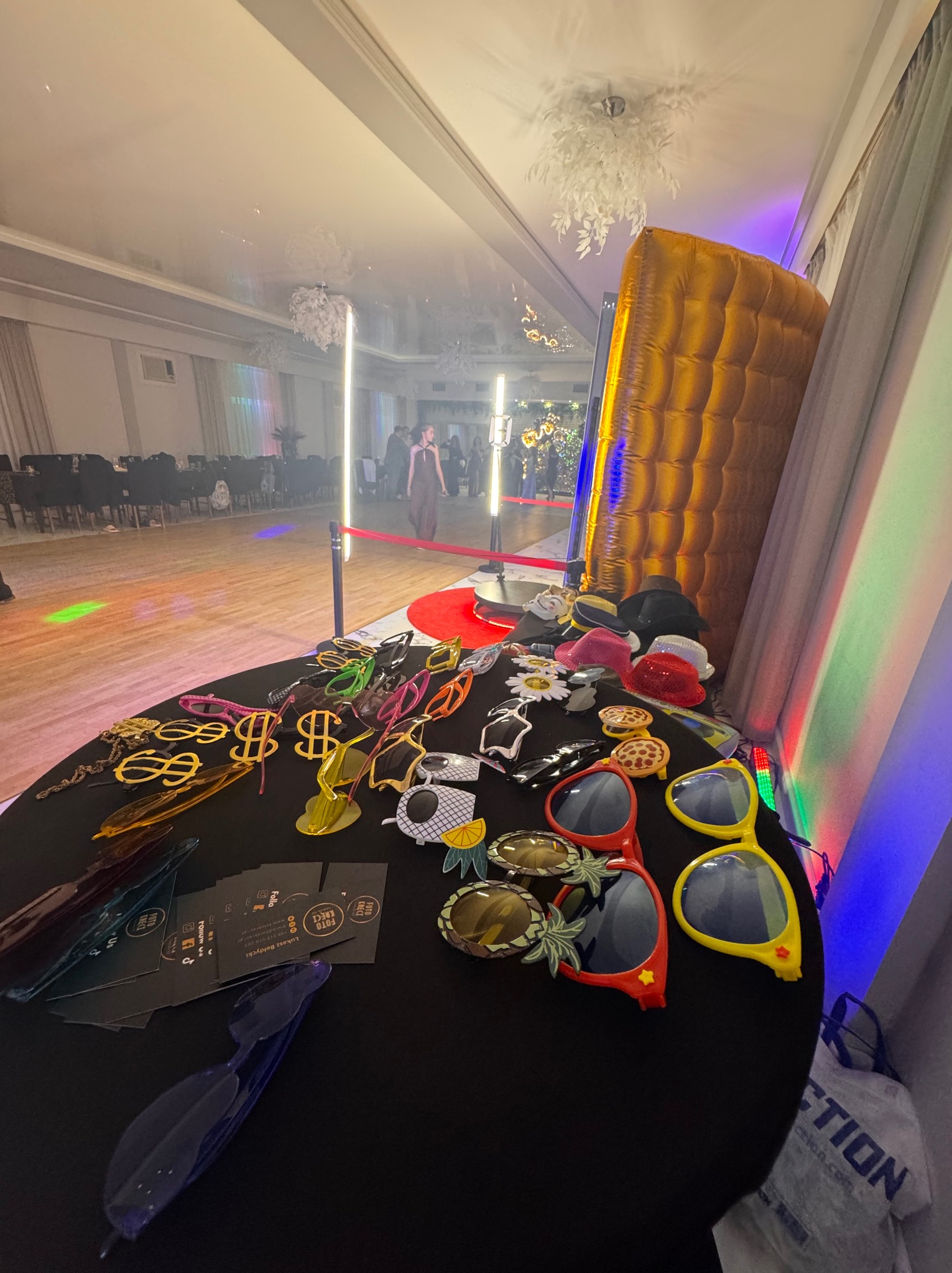 Stół z rekwizytami do fotobudki: okulary, kapelusze, symbole dolara. W tle dmuchana ścianka, oświetlenie i goście na sali balowej. Przygotowania do zabawy.