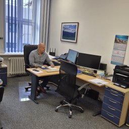 MARKA24 SPÓŁKA Z OGRANICZONĄ ODPOWIEDZIALNOŚCIĄ - Mężczyzna w biurze, siedzi przy biurku z dwoma monitorami, notuje. Na ścianie kalendarz i obrazek. Wnętrze biura z meblami i oknem z żaluzjami.