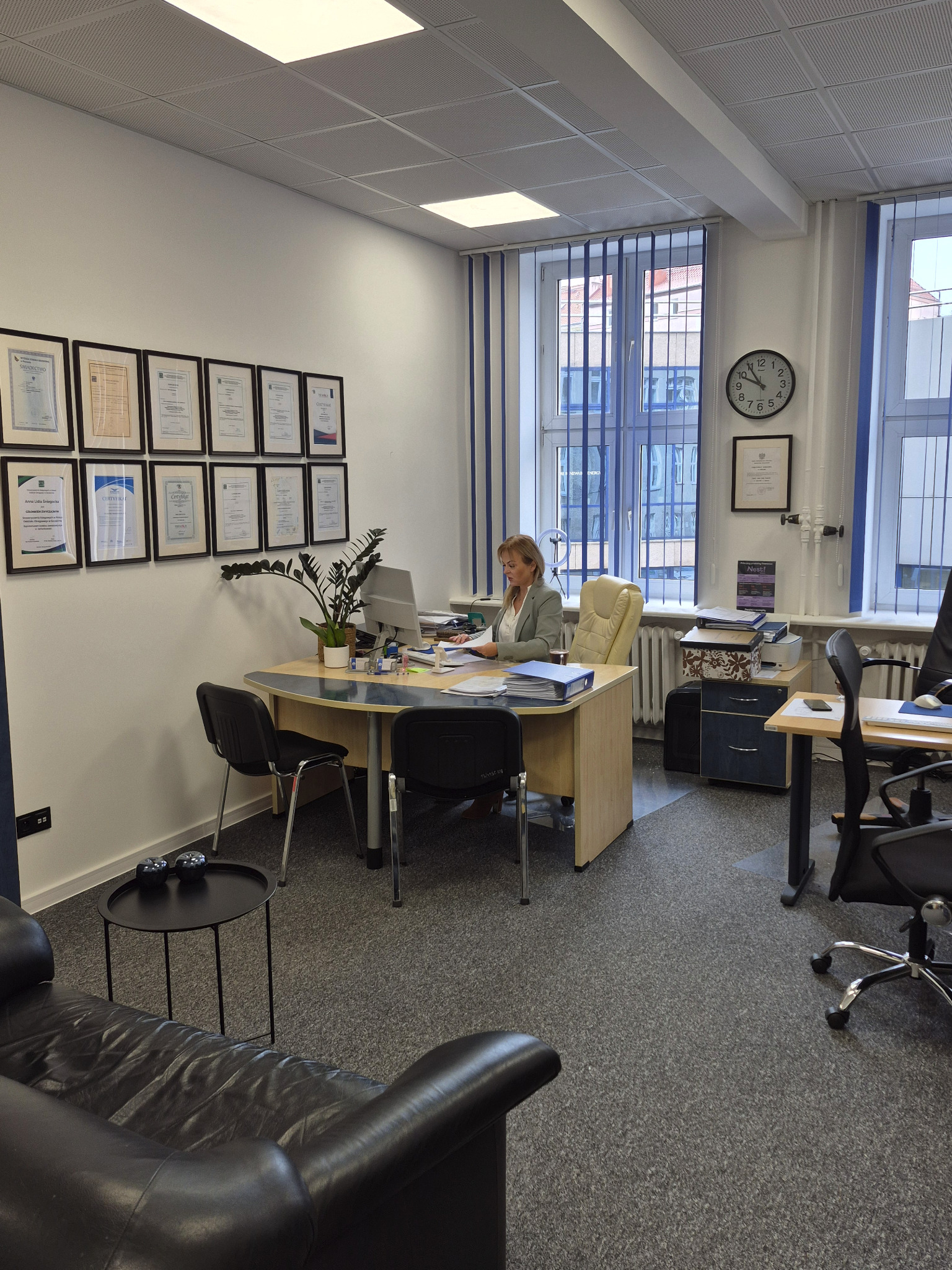Kobieta w biurze, siedząca przy biurku z komputerem i dokumentami. Na ścianie wiszą certyfikaty i dyplomy w ramkach, a w tle widać okno z żaluzjami.