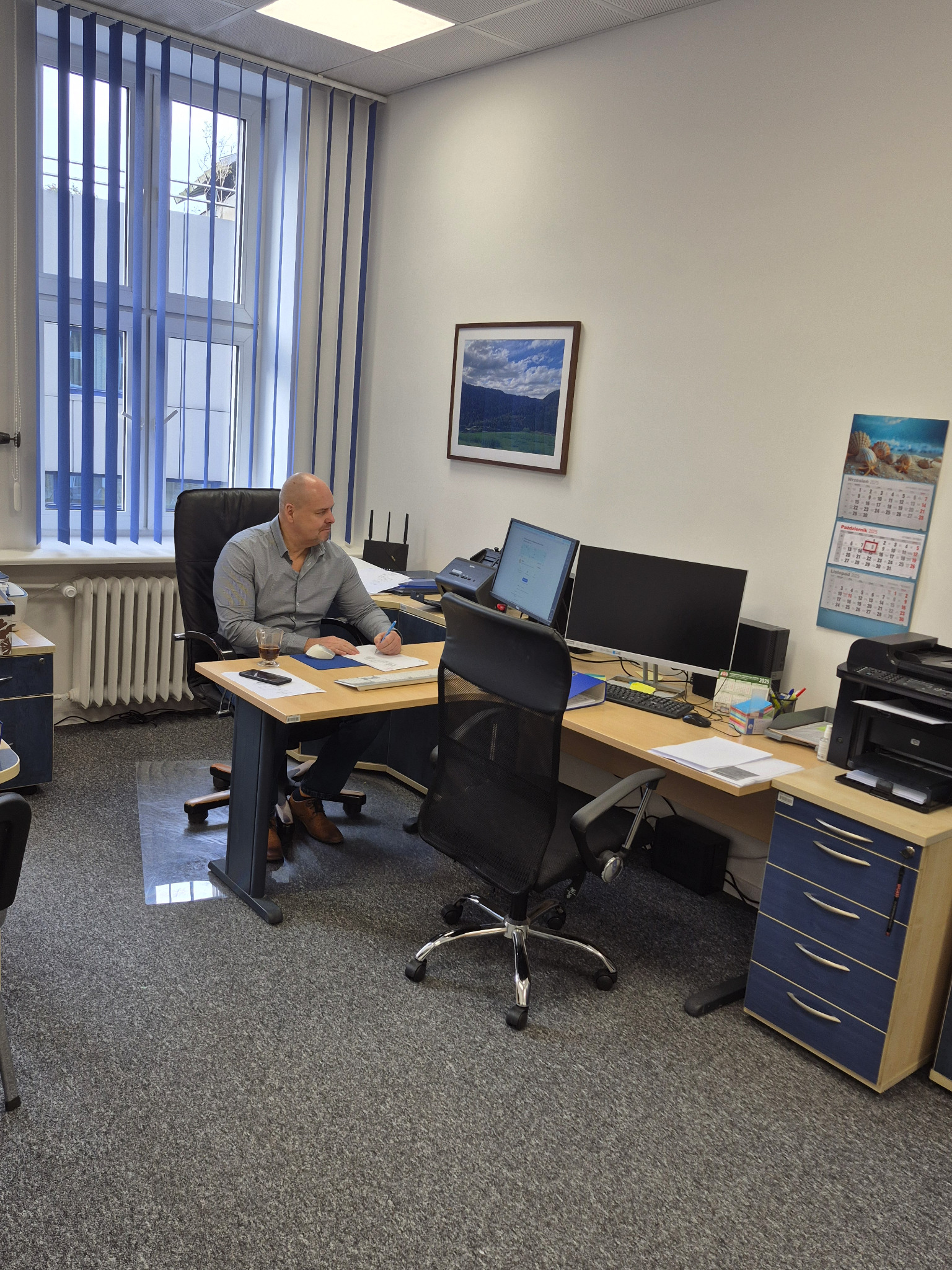 Mężczyzna w biurze, siedzi przy biurku z dwoma monitorami, notuje. Na ścianie kalendarz i obrazek. Wnętrze biura z meblami i oknem z żaluzjami.