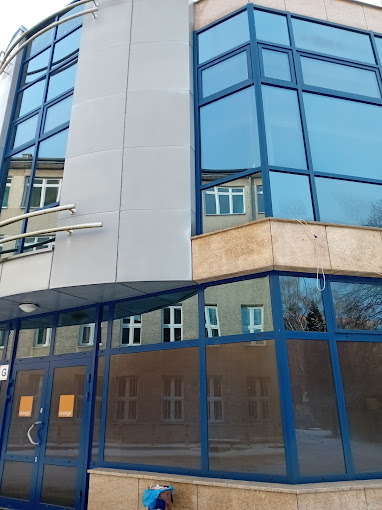 Nowoczesny budynek biurowy z niebieskimi ramami okiennymi i szarą elewacją. W oknach odbija się sąsiedni budynek, co dodaje głębi i złożoności kompozycji.