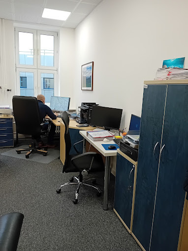 Wnętrze biura z pracownikiem siedzącym przy biurku, komputer, szafy biurowe. Na biurku dokumenty i akcesoria biurowe. Jasne pomieszczenie z oknem.