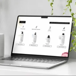 Laptop prezentuje stronę internetową z ofertą kosmetyków do pielęgnacji skóry trądzikowej. Widoczne cztery produkty z cenami i opcją dodania do koszyka. Minimalistyczny design.