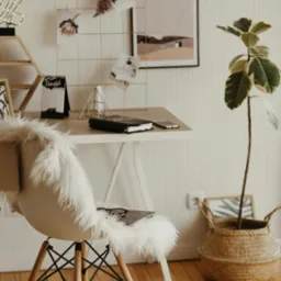 Jasne biuro z biurkiem, krzesłem z futrzanym siedziskiem, rośliną w koszu i tablicą inspiracji. Minimalistyczny design, ciepłe oświetlenie, parkiet w jodełkę.