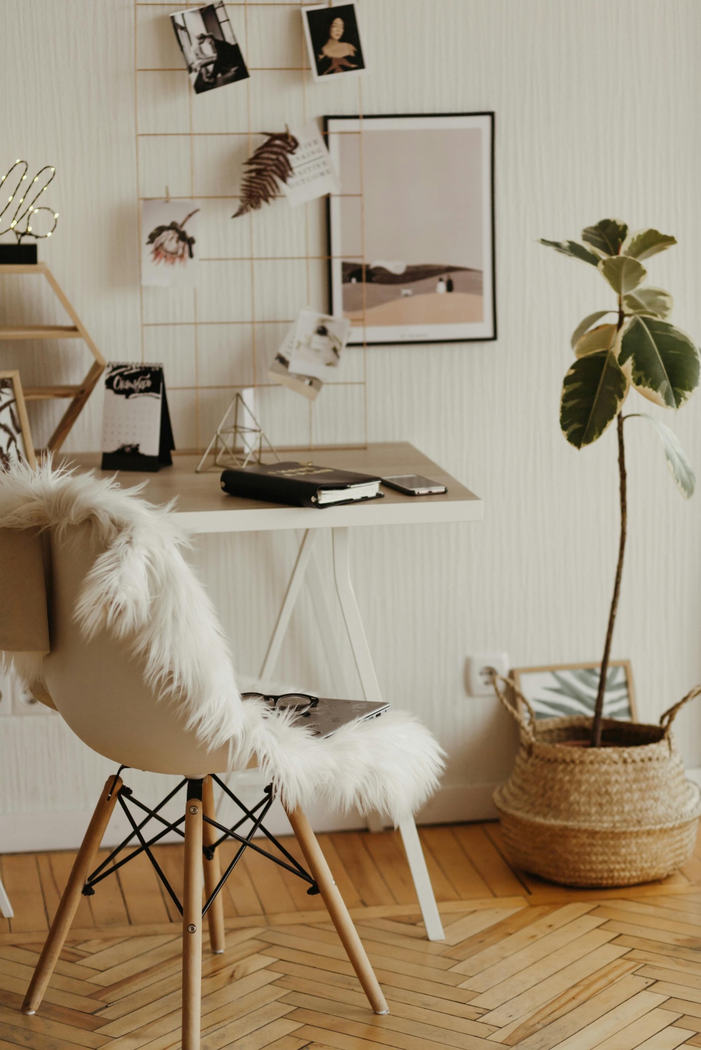 Jasne biuro z biurkiem, krzesłem z futrzanym siedziskiem, rośliną w koszu i tablicą inspiracji. Minimalistyczny design, ciepłe oświetlenie, parkiet w jodełkę.