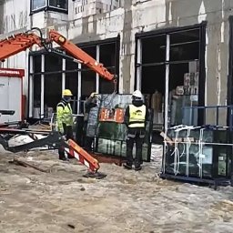 Eskaro - Montaż okien w surowym budynku za pomocą dźwigu. Pracownicy w kaskach i kamizelkach odblaskowych. Okna w czarnych ramach, budynek w trakcie budowy.