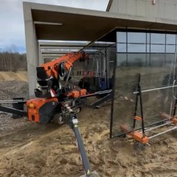 Eskaro - Kompaktowy żuraw gąsienicowy z pomarańczowym wysięgnikiem montuje duże przeszklenia w nowym budynku o surowej, betonowej konstrukcji.