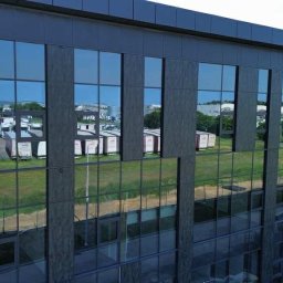 Eskaro - Elewacja biurowca z ciemnymi, lustrzanymi szybami, w których odbijają się przyczepy z logo firmy. Widok z zewnątrz, zieleń w tle.