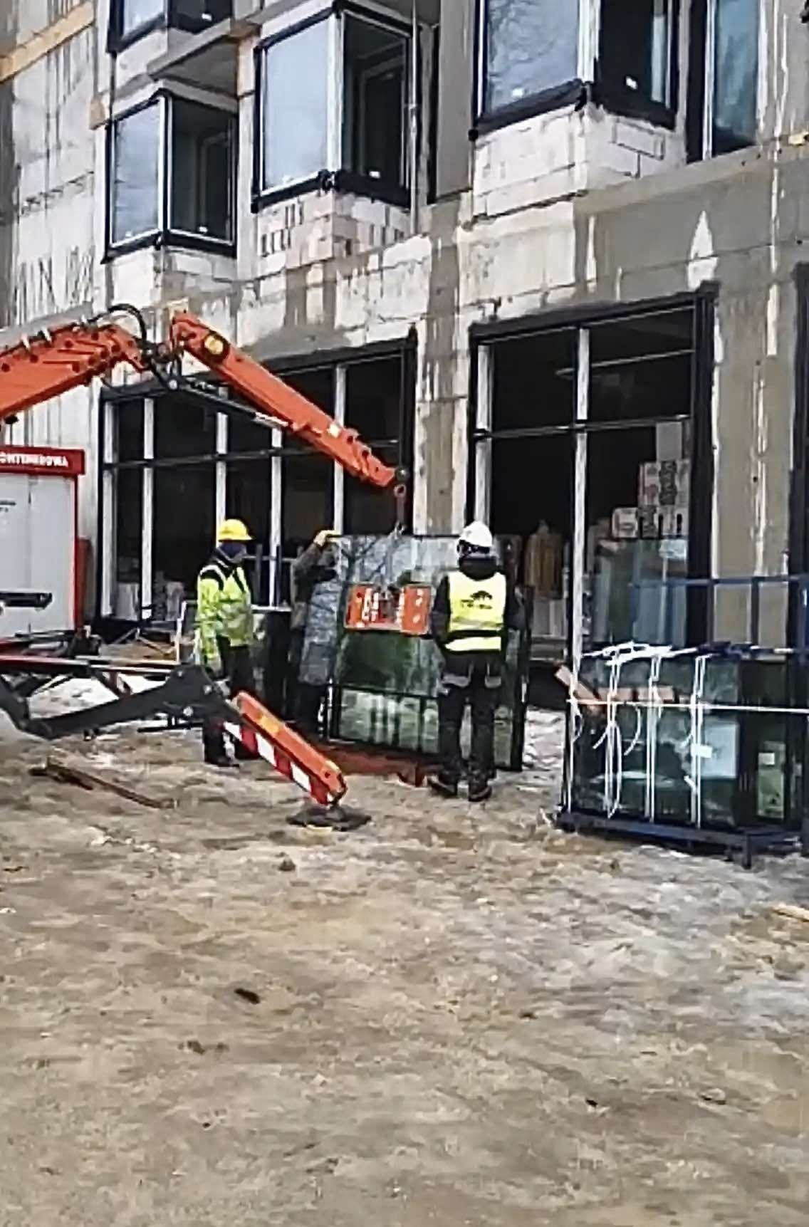 Montaż okien w surowym budynku za pomocą dźwigu. Pracownicy w kaskach i kamizelkach odblaskowych. Okna w czarnych ramach, budynek w trakcie budowy.