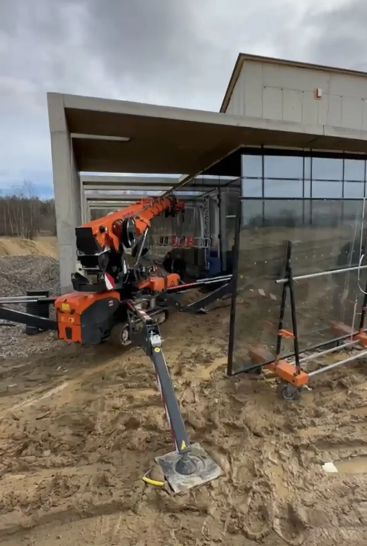 Kompaktowy żuraw gąsienicowy z pomarańczowym wysięgnikiem montuje duże przeszklenia w nowym budynku o surowej, betonowej konstrukcji.