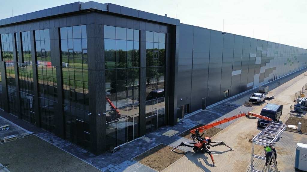 Nowoczesny budynek biurowy z szklaną fasadą i spider liftem, zaparkowane pojazdy, widok z góry na plac budowy z elementami rusztowania.