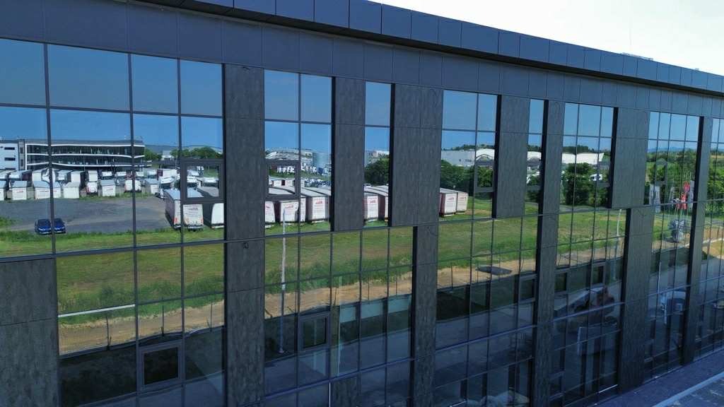 Elewacja biurowca z ciemnymi, lustrzanymi szybami, w których odbijają się przyczepy z logo firmy. Widok z zewnątrz, zieleń w tle.