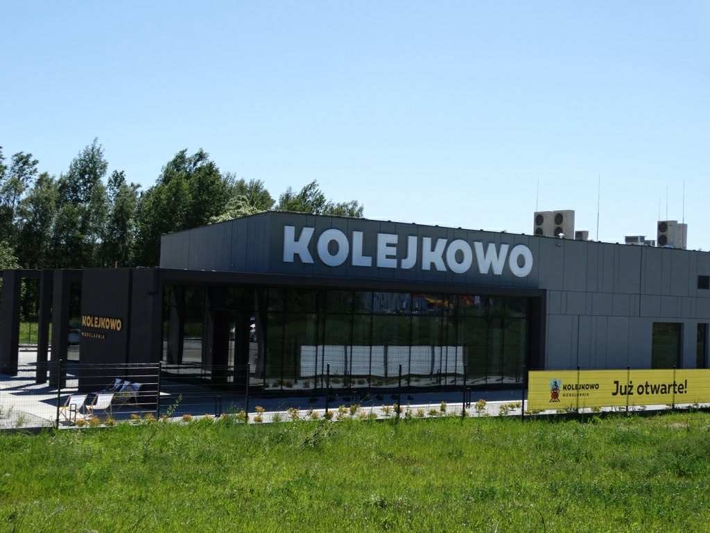 Nowoczesny budynek Kolejkowo z dużym napisem i banerem reklamowym 'Już otwarte!' na tle zieleni i błękitnego nieba w słoneczny dzień.