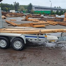 Nicolas Kurowiec - Przyczepa z ładunkiem drewna na błotnistym placu budowy. W tle składowane drewno i budynek magazynowy. Transport materiałów budowlanych.