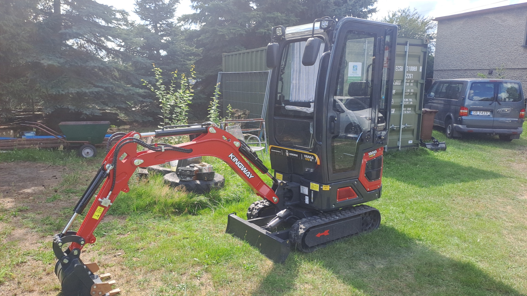 Czerwono-czarna mini koparka Kingway Yama HX10K na trawie, z kontenerem i samochodem w tle. Sprzęt budowlany, gotowy do pracy. Widoczne detale konstrukcji.