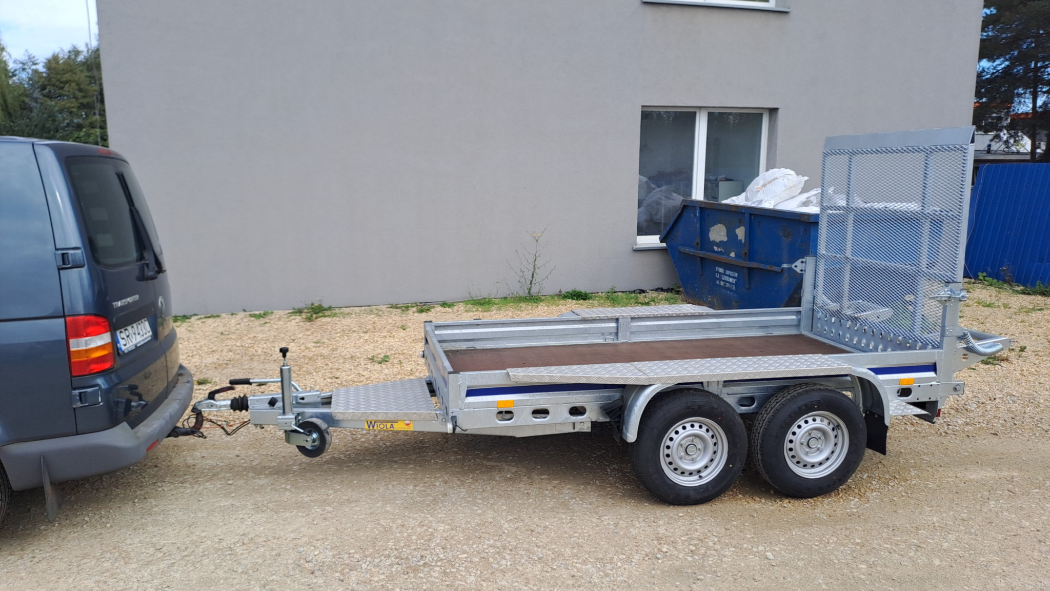 Przyczepa samochodowa marki Wiola podpięta do auta na żwirowym podłożu, w tle kontener na gruz i budynek. Gotowość do wywozu ziemi lub gruzu.