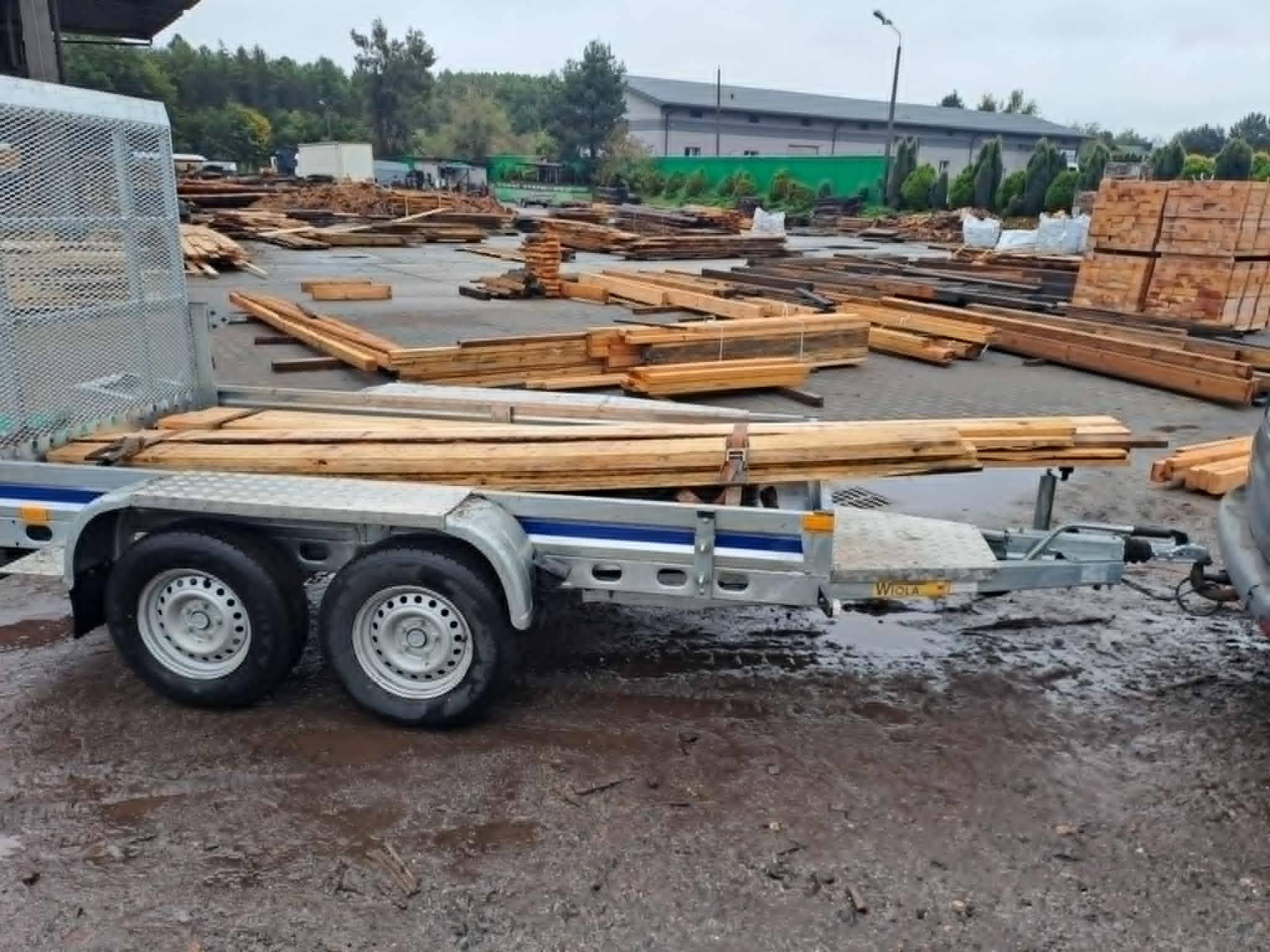 Przyczepa z ładunkiem drewna na błotnistym placu budowy. W tle składowane drewno i budynek magazynowy. Transport materiałów budowlanych.