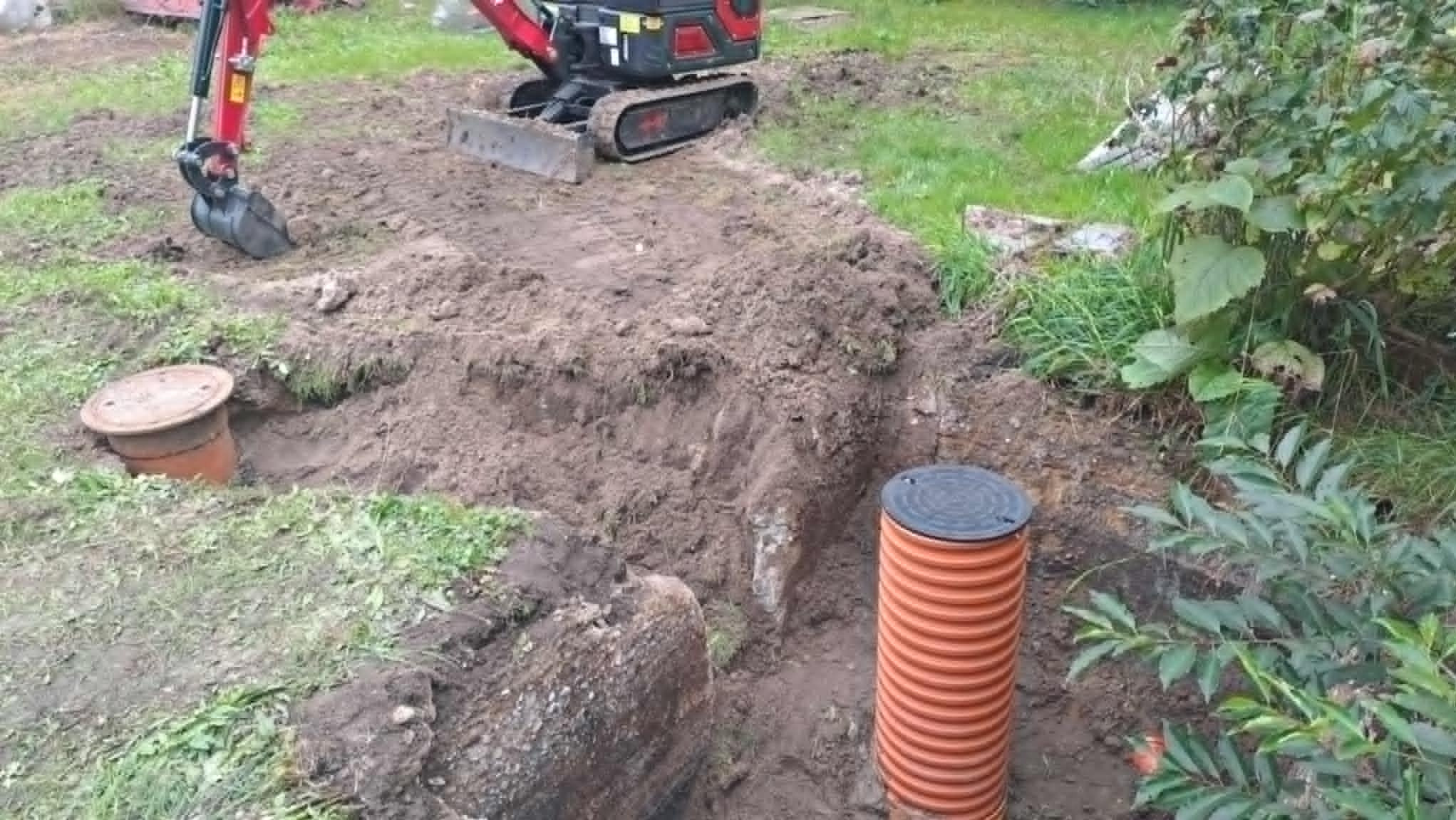 Mini koparka przy wykopie z rurami kanalizacyjnymi i studzienką. Prace ziemne w ogrodzie, widoczne świeżo wykopana ziemia i zieleń.