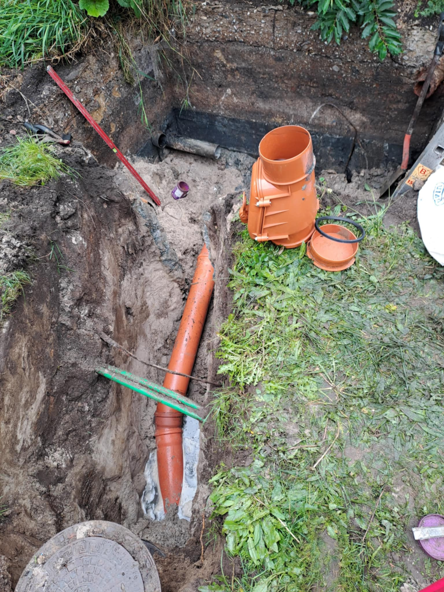 Wykop z pomarańczowymi rurami kanalizacyjnymi, poziomica i studzienka kanalizacyjna. Widoczne narzędzia i elementy instalacji w ziemi.