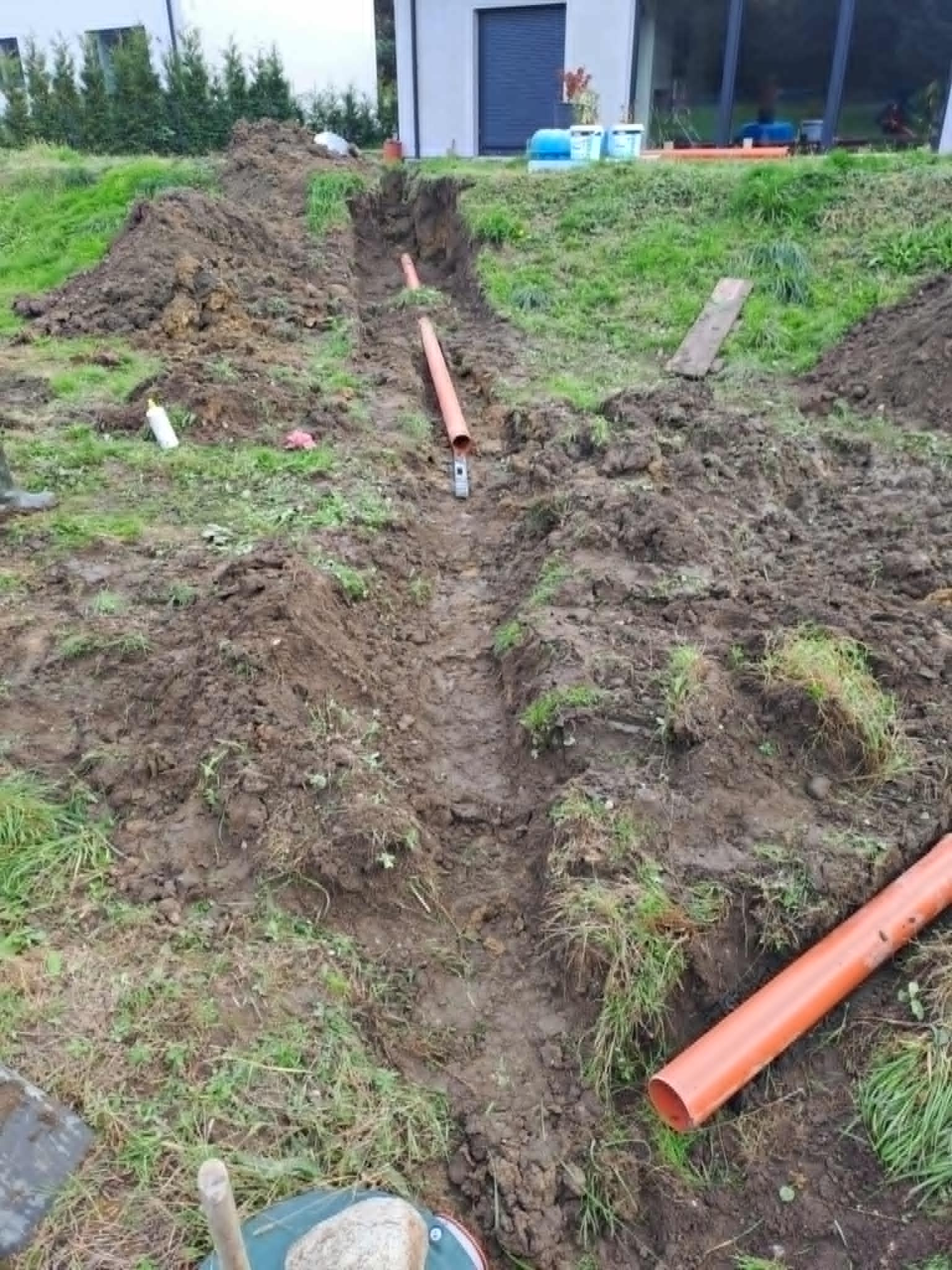 Wykopany wąski rów z pomarańczowymi rurami na dnie, widok z góry, ziemia i trawa po bokach, w tle budynek z dużymi oknami.