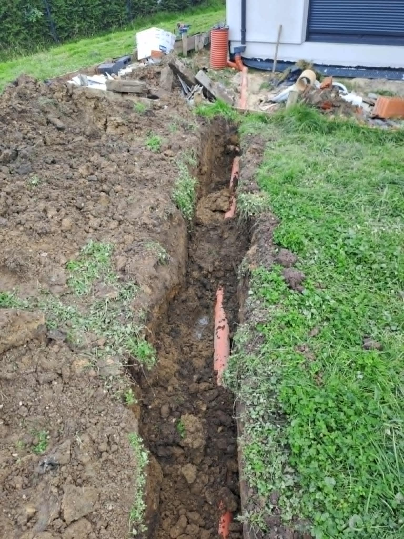 Wąski wykop pod rury kanalizacyjne na trawiastym terenie, widoczne pomarańczowe rury w ziemi. W tle fragment budynku z szarą roletą i materiały budowlane.