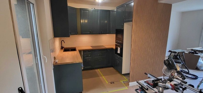 Granatowa zabudowa kuchenna z drewnianym blatem i panele ścienne z listewek. Widoczna piła do cięcia paneli. Ukończony etap prac remontowych.