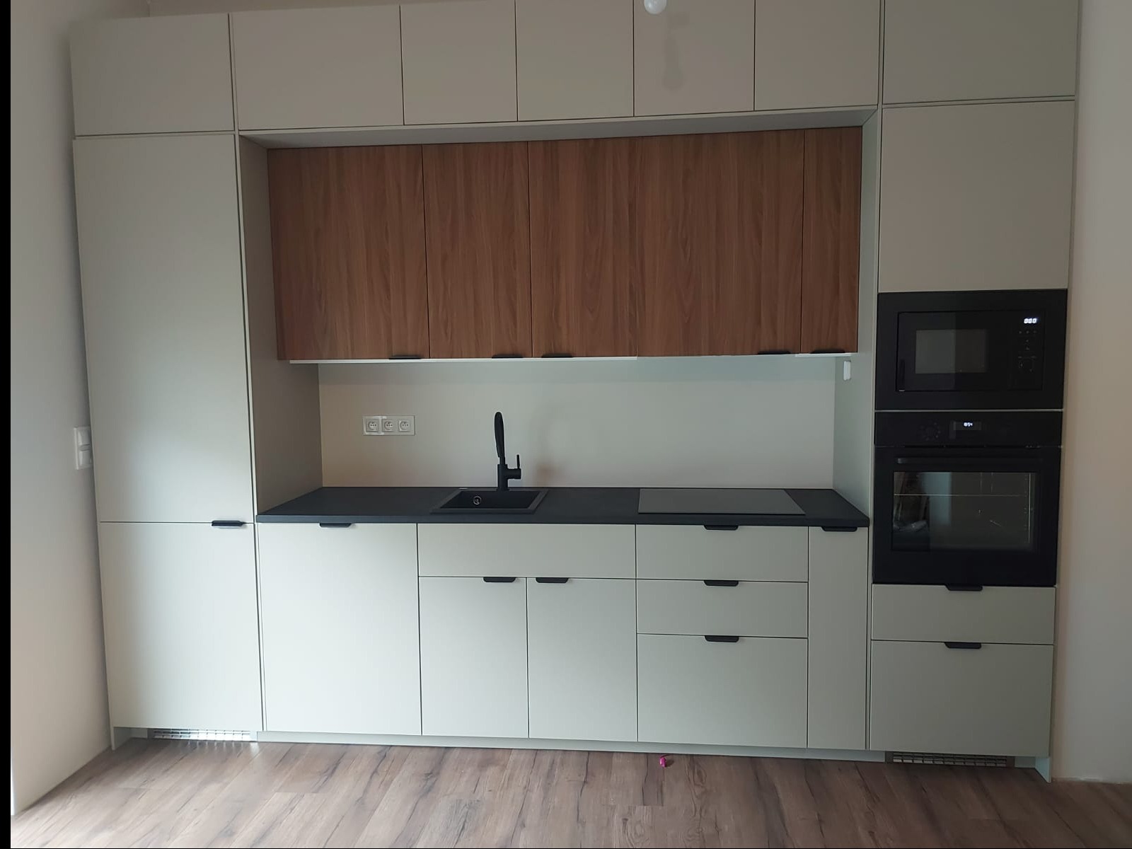 Nowoczesna kuchnia w odcieniach beżu i drewna, z czarnym blatem i sprzętem AGD. Minimalistyczny design, gładkie fronty szafek, drewniane akcenty nad blatem.