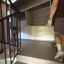 Expressfix - Przenoszenie szafy po schodach w bloku. Widoczny fragment klatki schodowej z metalową barierką i płytkami. Osoba w rękawicach niesie element mebla.