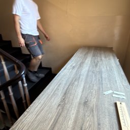 Expressfix - Pracownik transportuje panele podłogowe po schodach w budynku, używając miarki. Widoczne fragmenty schodów i ściany.