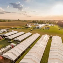 Widok z lotu ptaka na rozległy teren z rozstawionymi białymi namiotami eventowymi, przyczepami i pojazdami, w tle pola i lasy, oświetlone zachodzącym słońcem.