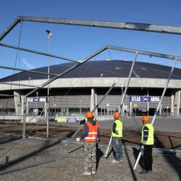 Krysiak Polska Nieruchomości - Montaż metalowej konstrukcji namiotu przed Atlas Areną. Czterech pracowników w kamizelkach i kaskach. Błękitne niebo w tle. Kąt ujęcia z dołu, widoczne elementy konstrukcyjne.