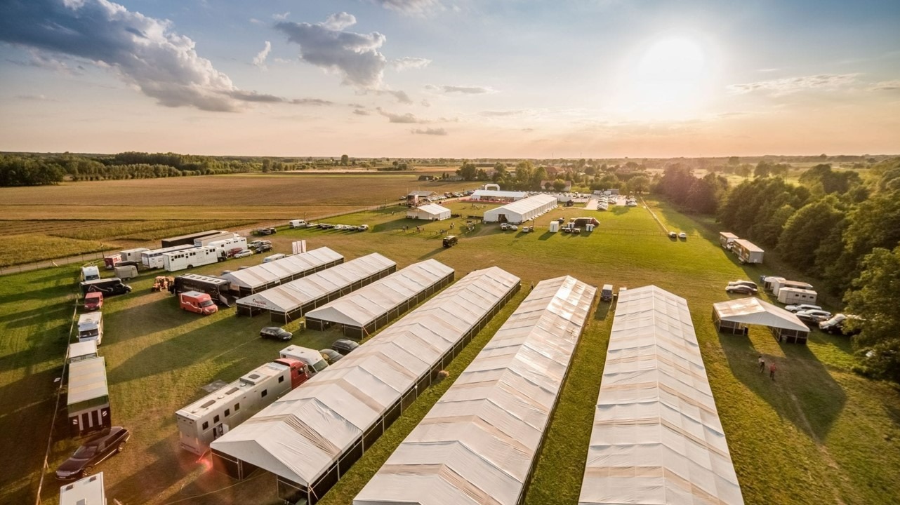 Widok z lotu ptaka na rozległy teren z rozstawionymi białymi namiotami eventowymi, przyczepami i pojazdami, w tle pola i lasy, oświetlone zachodzącym słońcem.