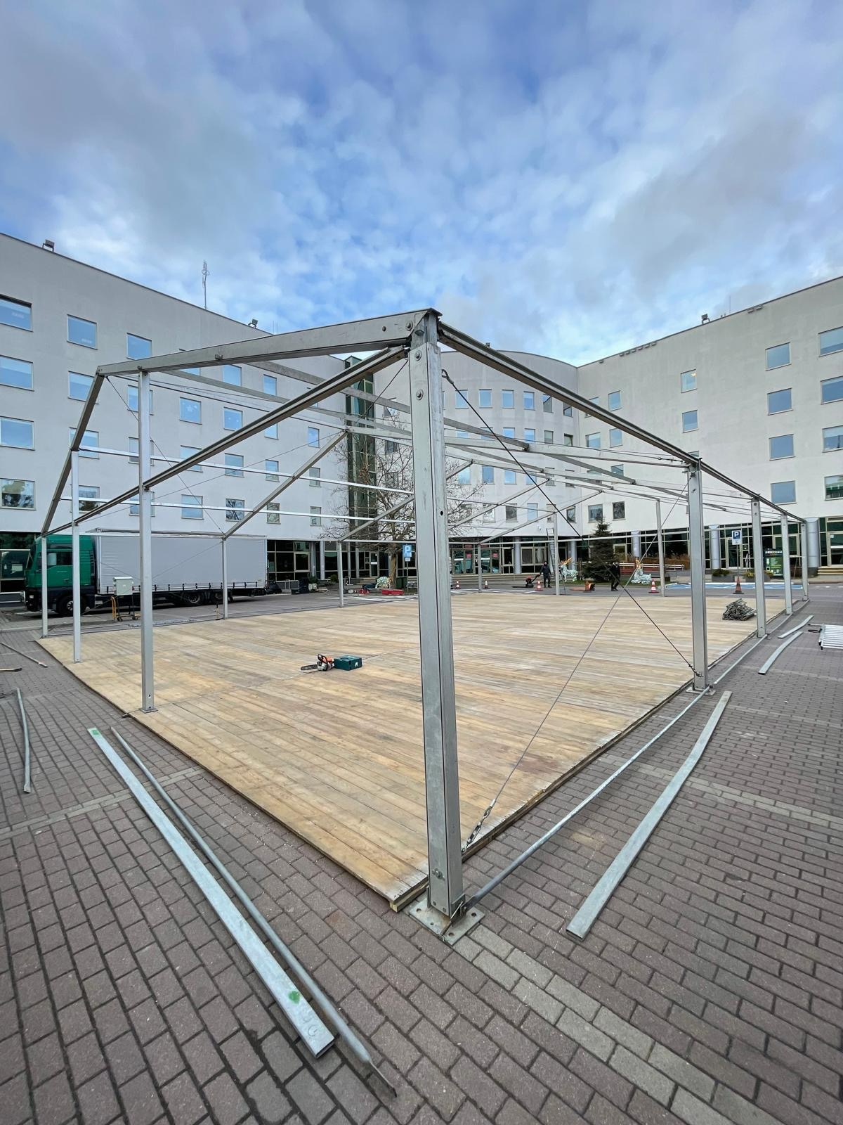 Konstrukcja metalowa namiotu z drewnianą podłogą na zewnątrz budynku. Widoczny sprzęt montażowy i elementy konstrukcyjne. Ujęcie szerokokątne.