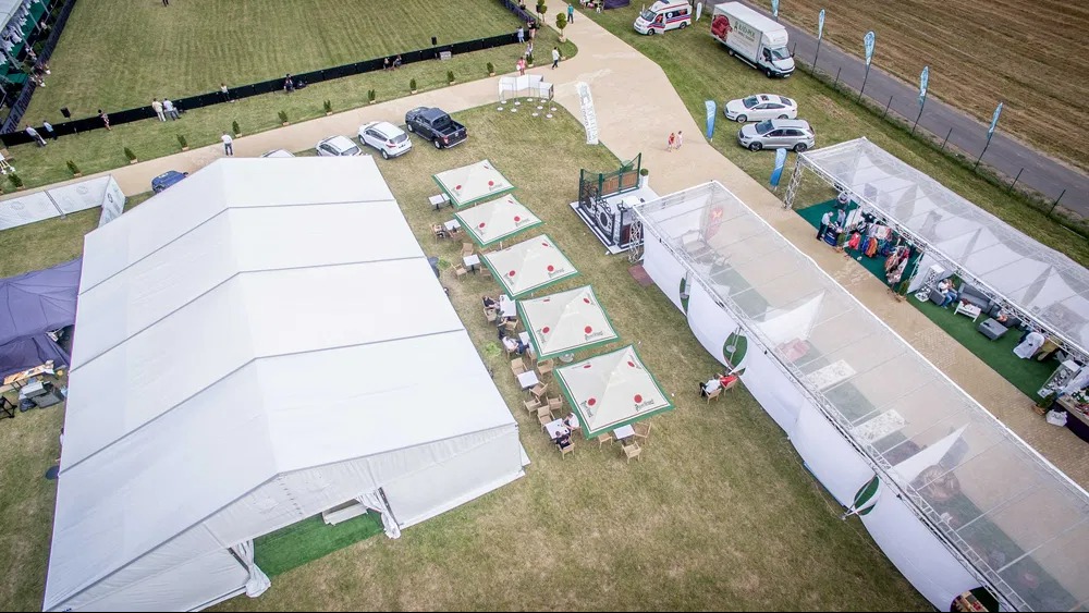 Widok z góry na rozległy teren eventowy z białymi namiotami, parasolami z logo, stolikami i krzesłami. Uczestnicy spacerują po terenie. W tle pojazdy i zieleń.