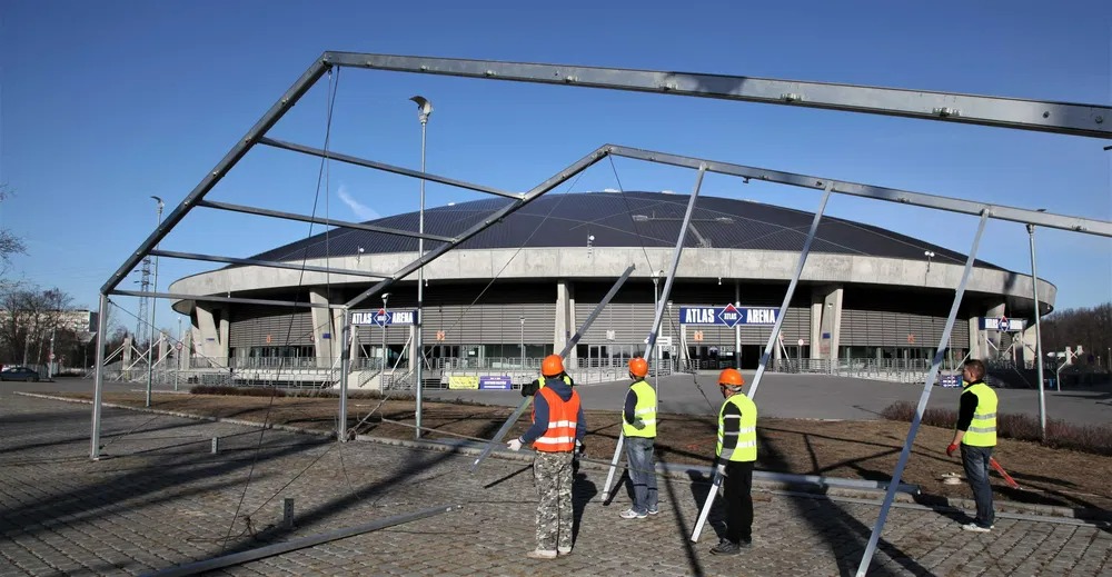 Montaż metalowej konstrukcji namiotu przed Atlas Areną. Czterech pracowników w kamizelkach i kaskach. Błękitne niebo w tle. Kąt ujęcia z dołu, widoczne elementy konstrukcyjne.